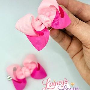 May include: Two hair bows made of ribbed fabric in shades of light pink and hot pink. Each bow features a central knot and layered loops. One bow is held up, while the other rests below, featuring a silver metal alligator clip on the back.