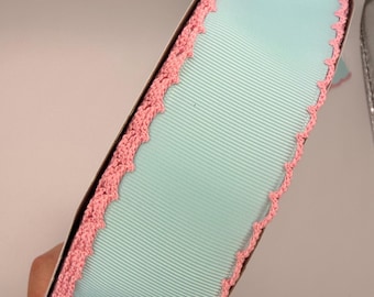 May include: A spool of light blue grosgrain ribbon featuring a decorative pink crocheted edge on both sides. The ribbon has a ribbed texture and is wrapped around a cardboard core, showcasing a consistent color palette and detailed trim.