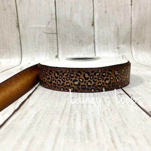 May include: A roll of brown ribbon with a leopard print design. The ribbon is on a white spool and has a gold glitter pattern. The text "Lainey's Loops" is visible on the ribbon.