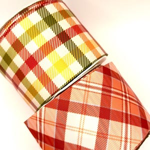 May include: Two spools of wide ribbon featuring plaid patterns. The top spool displays a grid of olive green, yellow, and red lines on a white background. The bottom spool features a pattern of intersecting red, orange, and white diagonal lines.