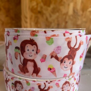 May include: White grosgrain ribbon featuring a repeating pattern of cartoon monkeys and ice cream cones. The monkeys are brown with pink accents, and the ice cream cones have various pastel colors. The ribbon is rolled up.
