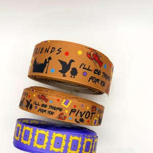 May include: Two rolls of brown ribbon featuring black silhouettes of a lobster, a duck, a chick, and the text "FRIENDS" and "I'LL BE THERE FOR YOU" and "PIVOT," plus one roll of purple ribbon with a repeating yellow picture frame pattern.