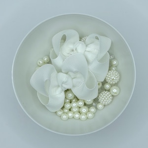 May include: A white bowl containing two white grosgrain ribbon bows and a collection of white spherical beads of varying sizes, including smooth round beads and textured, bumpy round beads, all arranged together in the center of the bowl.