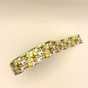 May include: A roll of white grosgrain ribbon featuring a repeating pattern of yellow bows with pink accents and small pencil tips. The ribbon has thin horizontal blue lines running across the white background, resembling notebook paper.