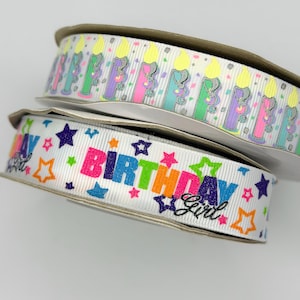 May include: Two spools of white grosgrain ribbon. The top spool features a repeating pattern of colorful birthday candles with yellow flames. The bottom spool displays the text "Birthday Girl" in multicolored glittery letters with stars.