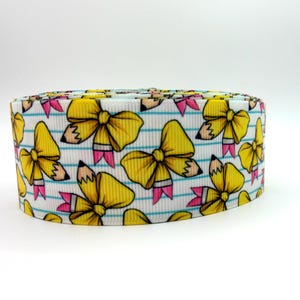 May include: A roll of white grosgrain ribbon featuring a repeating pattern of yellow bows shaped like pencils with pink erasers and black tips, set against a background of thin horizontal blue lines resembling notebook paper.