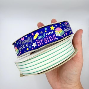 May include: Two spools of grosgrain ribbon. The top spool is royal blue with a repeating pattern of yellow pencils, colorful stars, stacks of books, and the text "Beauty and Brains". The bottom spool is white with thin, horizontal green stripes.