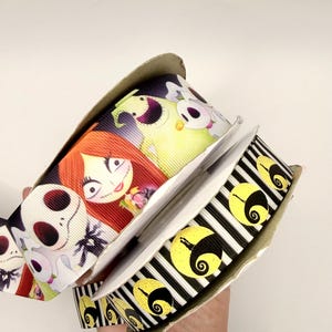 May include: Two spools of grosgrain ribbon. One spool features a repeating pattern of characters with red hair, white faces, and a pale green creature. The second spool features a black and white striped pattern with yellow circular moon icons.