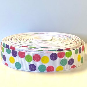 May include: A spool of white grosgrain ribbon featuring a repeating pattern of glittery polka dots in shades of purple, pink, yellow, and light green. The ribbon is wound into a tight, circular coil, displaying the colorful dotted texture.