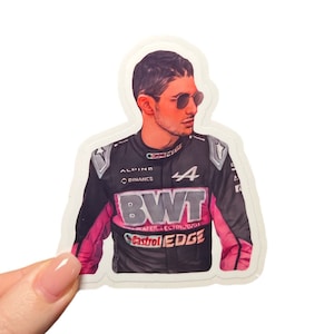 Op de afbeelding: Een sticker met een raceautocoureur die een zwart en roze racepak draagt met de logo's "Alpine", "Binance", "BWT" en "Castrol EDGE". De coureur draagt een zonnebril en heeft een vastberaden uitdrukking op zijn gezicht.
