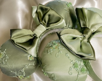 May include: A pair of green satin and lace Minnie Mouse ears with green satin bows. The ears are decorated with green sequins and beads.
