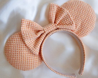 May include: A pair of orange and white checkered fabric mouse ears with a matching bow. The ears are attached to a white headband.
