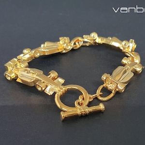 Könnte beinhalten: Ein goldfarbenes Armband mit einer Reihe von Miniatur-Rennwagen-Anhängern. Das Armband hat einen Knebelverschluss und wird auf einer dunklen Oberfläche präsentiert. Der Markenname "vanbelle" ist oben rechts sichtbar.