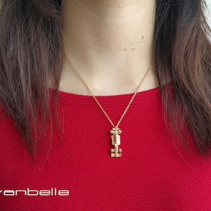 May include: Gold-coloured necklace featuring a race car pendant suspended from a fine chain. The person in the image is wearing a red top. The brand name "vanbelle" is visible in the bottom left corner.
