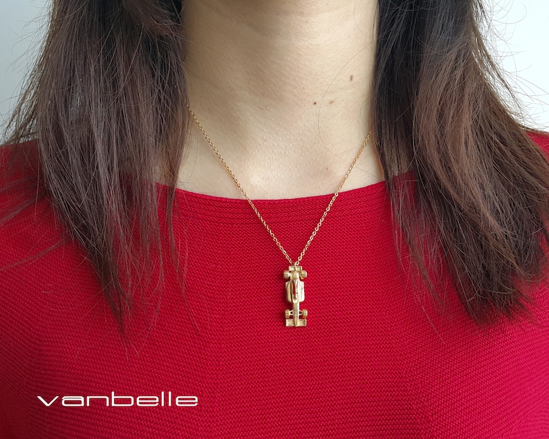 May include: Gold-coloured necklace featuring a race car pendant suspended from a fine chain. The person in the image is wearing a red top. The brand name "vanbelle" is visible in the bottom left corner.