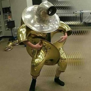 May include: A person wearing a gold-colored tuba costume, with the bell of the tuba forming a skirt and the mouthpiece forming a helmet. The person is playing the tuba with their right hand, and the tuba is making a blur effect.