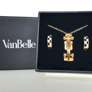 May include: A gold necklace and earring set with a race car pendant and hoop earrings with black and white chequered accents. The set is presented in a black box with the brand name "VanBelle" in white.