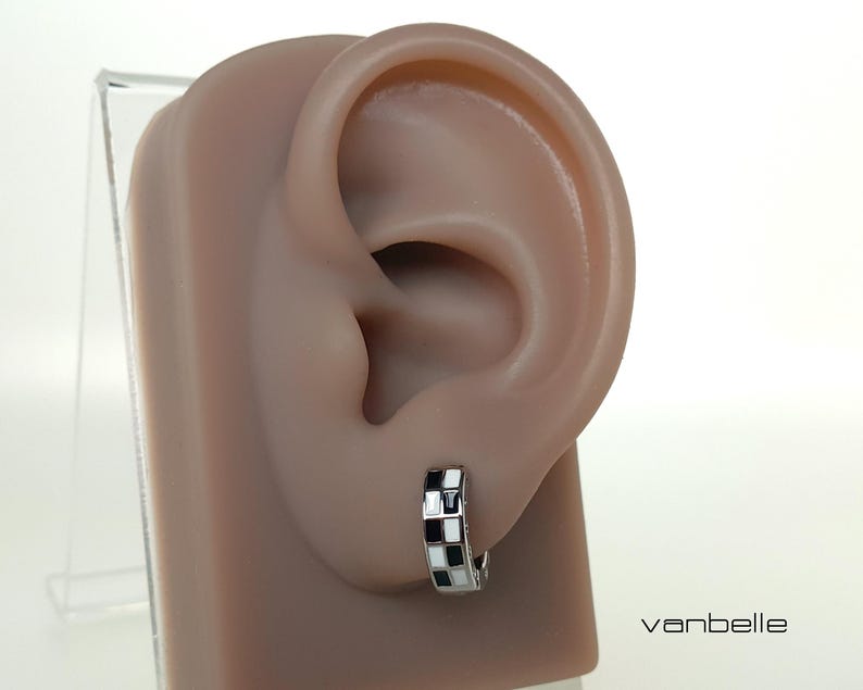 May include: A silver hoop earring with a black and white chequered pattern is displayed on a mannequin ear. The earring features a small, geometric design. The brand name "vanbelle" is visible in the lower right corner.