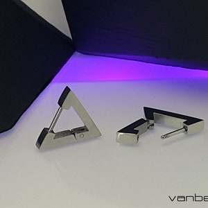 Puede incluir: Dos pendientes de plata en forma de triángulo con cierre de bisagra. Los pendientes están sobre una superficie blanca.