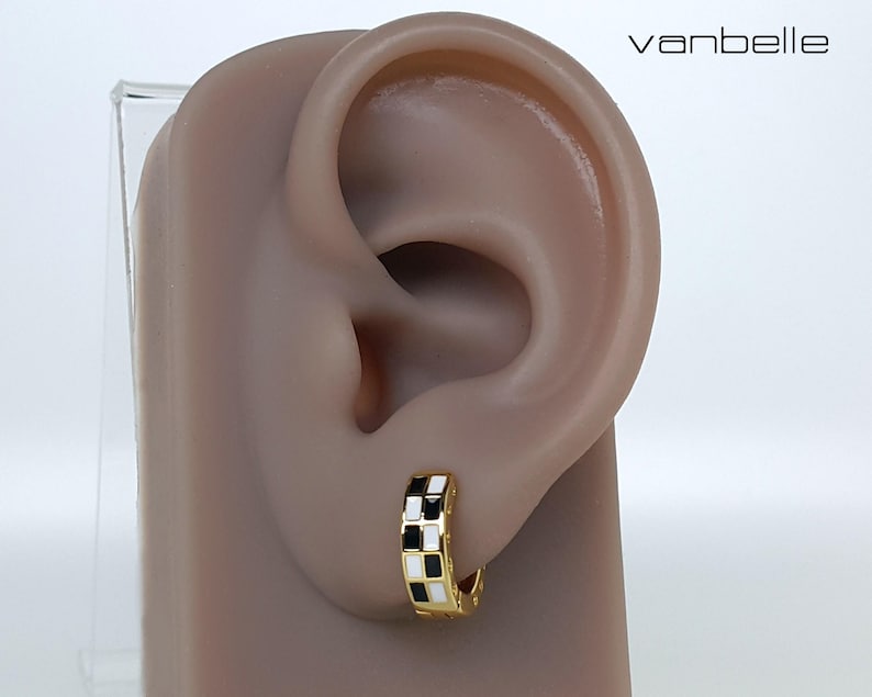 May include: Gold-tone hoop earring with a black and white chequered design. The earring is shown on a mannequin ear. The word "vanbelle" is in the upper right corner.