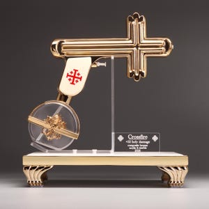 May include: A gold-toned sculpture shaped like a pistol with a cross-shaped barrel, featuring a red Jerusalem cross emblem on the handle. A clear circular chamber contains a gold-toned figure, mounted on a rectangular base with a plaque reading Crossfire +33 holy damage composite bronze acrylic & marble 2026.