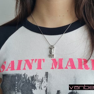 May include: Silver pendant necklace with a geometric design. The pendant hangs from a fine chain. The model is wearing a white ribbed t-shirt with black sleeves and the text "SAINT MARKS" in pink.