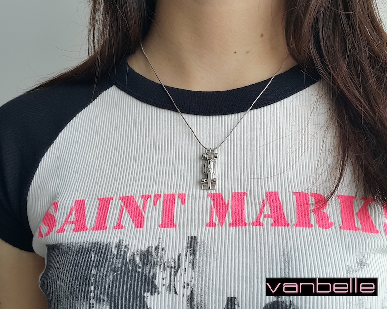 May include: Silver pendant necklace with a geometric design. The pendant hangs from a fine chain. The model is wearing a white ribbed t-shirt with black sleeves and the text "SAINT MARKS" in pink.