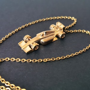 May include: A gold-coloured pendant necklace with a Formula 1 race car charm. The charm is a detailed miniature of a race car, suspended from a delicate gold chain. The necklace is displayed on a dark surface, highlighting the gold colour.