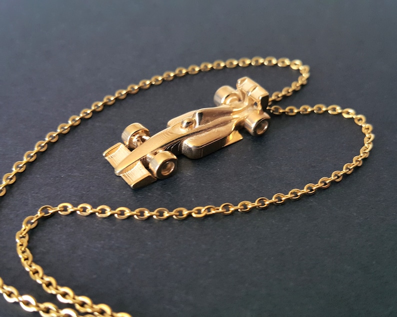 May include: A gold-coloured pendant necklace with a Formula 1 race car charm. The charm is a detailed miniature of a race car, suspended from a delicate gold chain. The necklace is displayed on a dark surface, highlighting the gold colour.