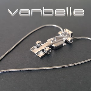 May include: A silver pendant necklace with a miniature race car charm. The charm has detailed wheels and aerodynamic features. The necklace chain forms a heart shape. The word "vanbelle" is displayed in white text.