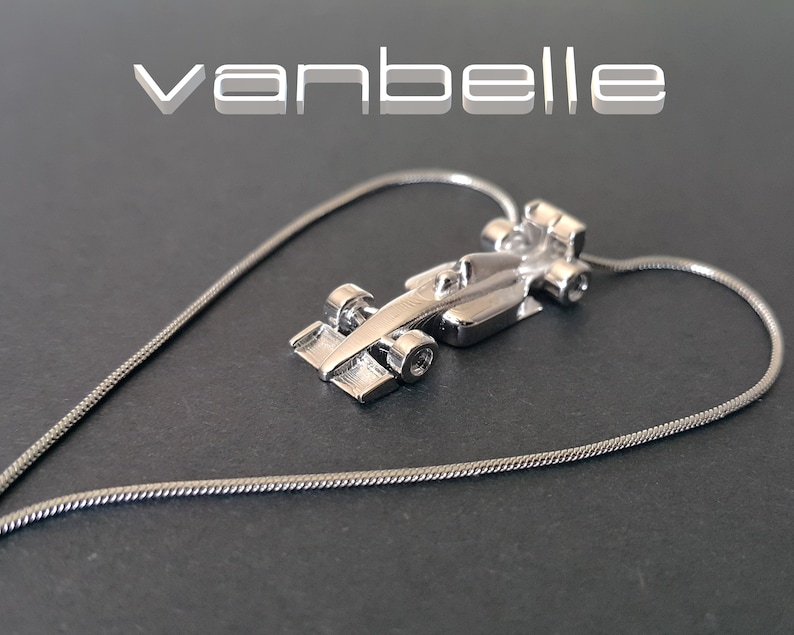 May include: A silver pendant necklace with a miniature race car charm. The charm has detailed wheels and aerodynamic features. The necklace chain forms a heart shape. The word "vanbelle" is displayed in white text.