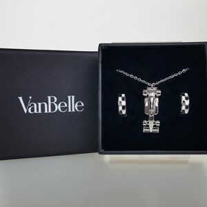 May include: A silver necklace and earrings set presented in a black box. The necklace has a race car pendant, and the earrings feature a chequered pattern. The box is open, displaying the jewellery. The brand name "VanBelle" is visible on the box.