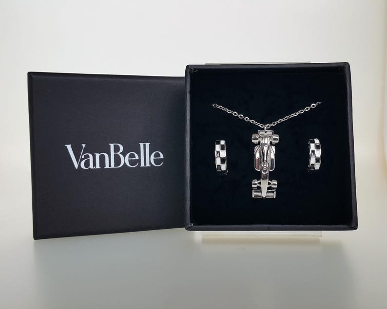 May include: A silver necklace and earrings set presented in a black box. The necklace has a race car pendant, and the earrings feature a chequered pattern. The box is open, displaying the jewellery. The brand name "VanBelle" is visible on the box.