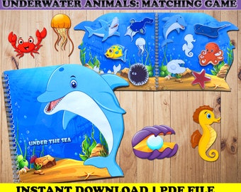 May include: A matching game featuring underwater animals. The game includes a spiral-bound book with a blue dolphin on the cover and the text "UNDER THE SEA". Various sea creatures like a crab, jellyfish, shark, and seahorse are displayed. The text "UNDERWATER ANIMALS: MATCHING GAME" is at the top.