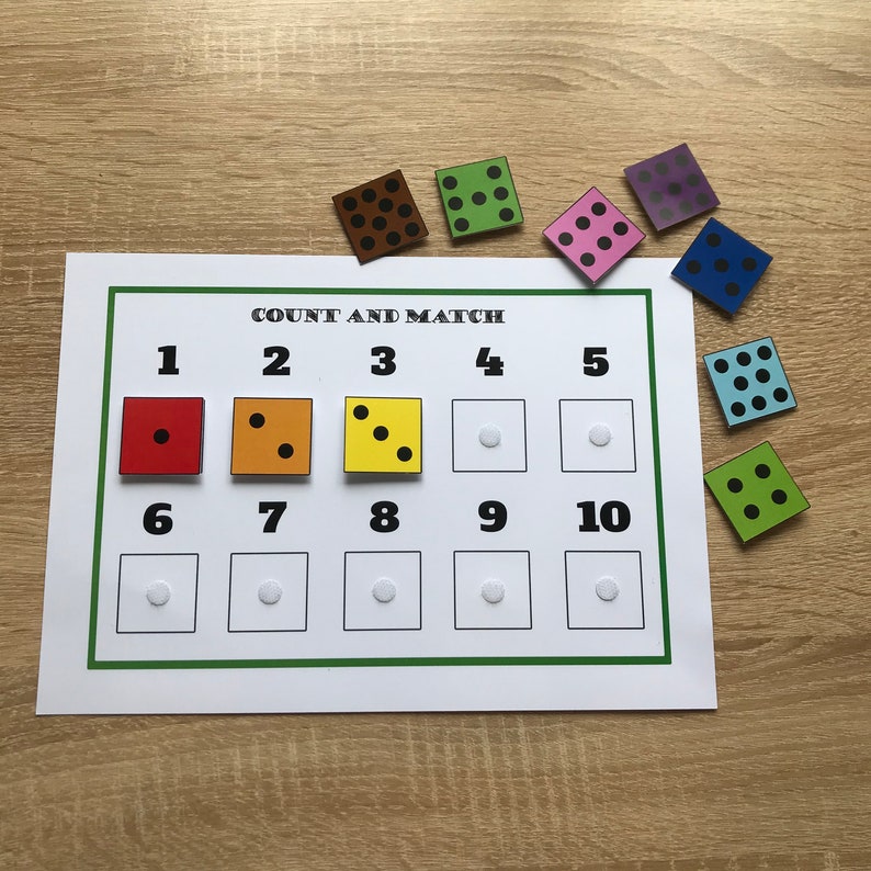 K&ouml;nnte beinhalten: Ein Z&auml;hlspiel mit wei&szlig;em Hintergrund und gr&uuml;nem Rand. Das Spiel beinhaltet ein bedrucktes Blatt mit den Zahlen 1-10 und passenden Quadraten. Bunte W&uuml;rfel mit schwarzen Punkten sind um das Blatt verstreut. Der Text "COUNT AND MATCH" steht oben.