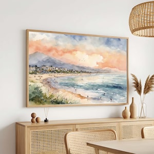 May include: A rectangular wall art print featuring a watercolor landscape of a coastal beach scene. The painting depicts a sandy shoreline, blue ocean waves, and a town nestled against mountains under a soft pink and orange sunset sky.