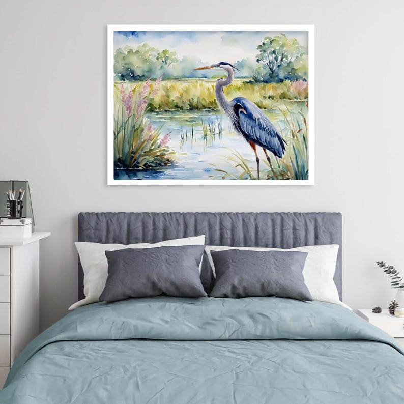 May include: A framed watercolor painting of a blue heron standing in a marshy wetland. The bird has blue and grey feathers with a long neck and pointed beak. The background features soft green trees, tall grasses, and pink wildflowers.