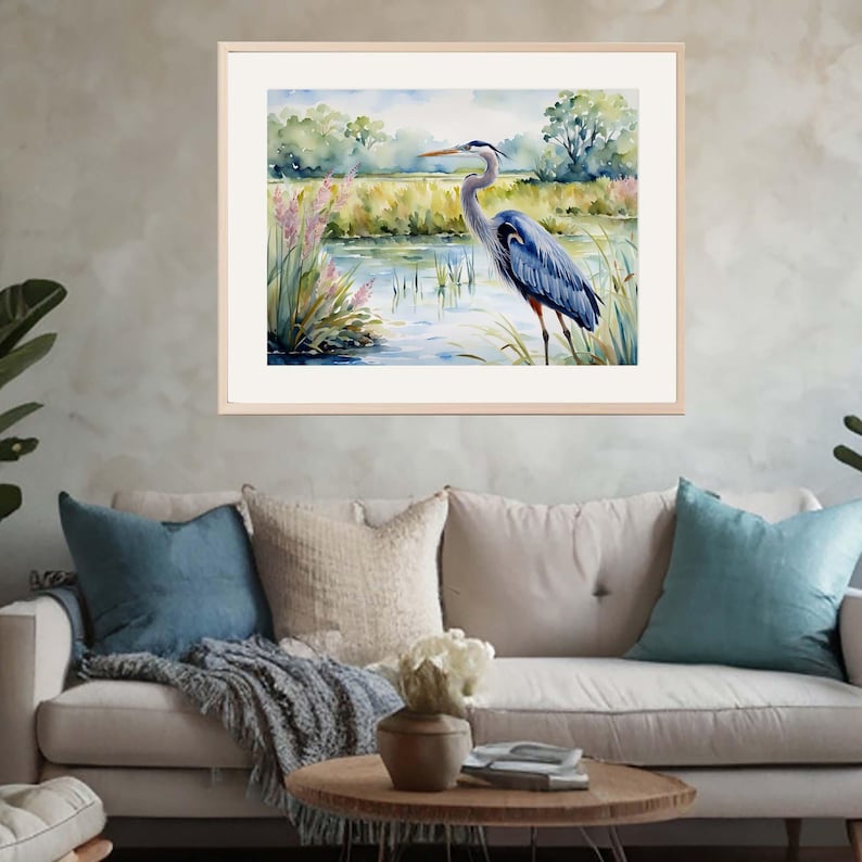 May include: A framed watercolor painting depicting a great blue heron standing in a marshy wetland. The bird features blue-gray plumage and a long neck, positioned near tall grasses and water with soft green and blue tones in the background.