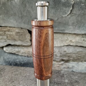 May include: A wooden pepper grinder with a stainless steel top and bottom. The grinder is sitting on a gray surface with black peppercorns scattered around it.