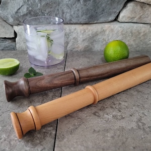 May include: Two wooden muddlers, one brown and one light wood, with a glass of ice water and a lime wedge in the background.