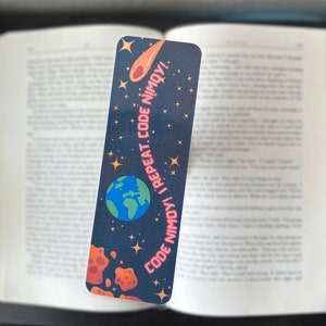 May include: A rectangular bookmark with a dark blue background featuring a cartoon Earth, a comet, and stars. The text "CODE NIMOY! I REPEAT. CODE NIMOY!" is printed in pink. The bookmark is placed in an open book.