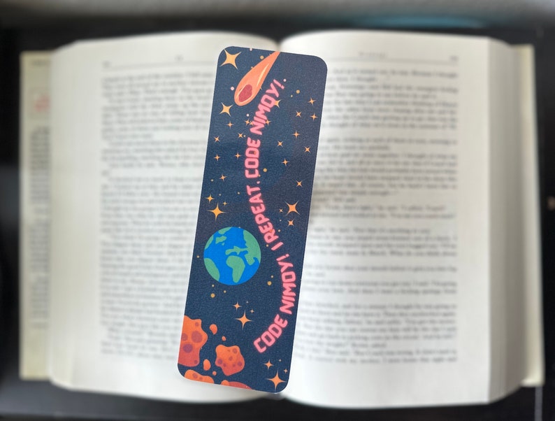 Pu&ograve; includere: Un segnalibro rettangolare con sfondo blu scuro con una Terra dei cartoni animati, una cometa e stelle. Il testo "CODE NIMOY! I REPEAT. CODE NIMOY!" &egrave; stampato in rosa. Il segnalibro &egrave; posto in un libro aperto.