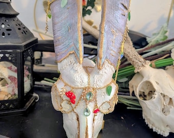 May include: A decorative goat skull with painted horns and gold accents. The horns are painted in shades of purple and gold, adorned with gold chains and colorful gemstone charms. The skull is decorated with red and green mushroom charms.