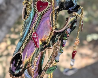 May include: A decorative skull with iridescent green, purple, and black paint. Gold chains and charms, including a red heart, coffin, and colorful beads, hang from the skull. A large, faceted crystal is attached to the jaw.