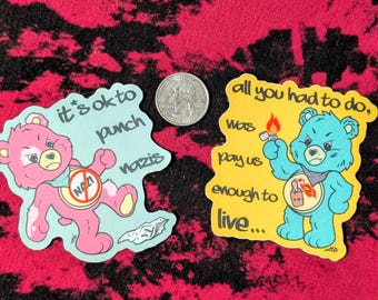 May include: Two die-cut vinyl stickers featuring cartoon bears. The pink bear sticker reads "it's ok to punch nazis" with a "no nazi" symbol. The yellow bear sticker reads "all you had to do, was pay us enough to live..." with a lighter. A US quarter is shown for scale.
