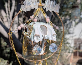 May include: A handcrafted, celestial-themed hanging ornament. It features a cat skull painted white, adorned with clear quartz crystals, pink beads, and gold-colored chains. A wooden moth silhouette hangs below, creating a unique, mystical aesthetic.
