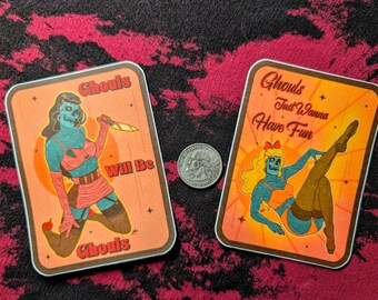 May include: Two rectangular stickers featuring blue-skinned skull-faced figures in pin-up style poses. The left sticker reads Ghouls Will Be Ghouls and shows a figure holding a knife. The right sticker reads Ghouls Just Wanna Have Fun. A US quarter is centered between them.