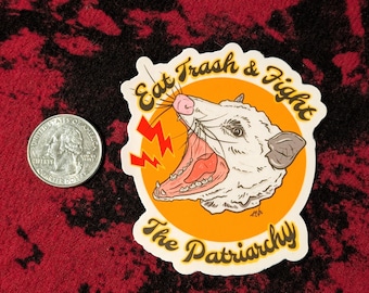 May include: A die-cut sticker featuring an illustration of a white opossum with its mouth open, set against a circular orange background. The text 'Eat Trash & Fight The Patriarchy' is written in black. A US quarter is shown for scale.