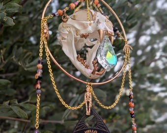 May include: A decorative hanging ornament featuring a cat skull, a large iridescent crystal, and a black planchette with the words "HELLO" and "GOODBYE". The ornament is adorned with gold chains, beads, and a gold ring.