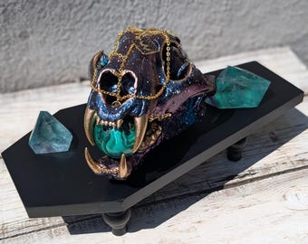 May include: A decorative, iridescent skull with gold accents and a malachite orb in its mouth, resting on a black platform. Two teal-colored crystals flank the skull. The skull is adorned with a gold chain.